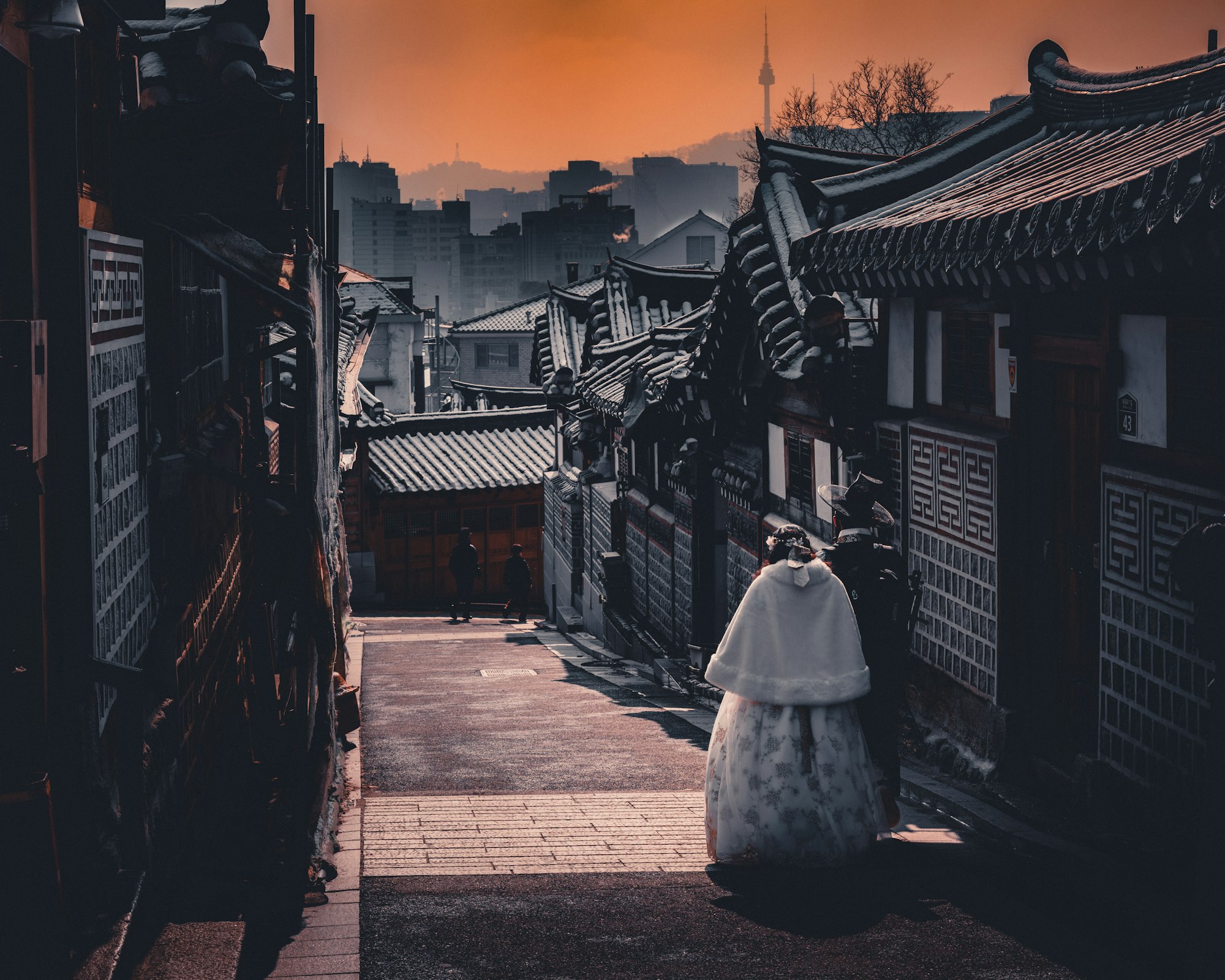 A Bukchon hanok alley at dusk with tiled roofs, two visitors in hanbok and Namsan Tower behind